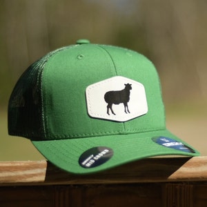 May include: A green trucker hat with a white patch featuring a black silhouette of a sheep. The hat has a mesh back and a mid-crown.