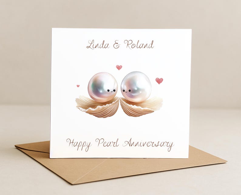 Op de afbeelding: Een witte wenskaart met de tekst "Linda & Roland" en "Happy Pearl Anniversary". De kaart toont twee cartoonparels met gezichten, rustend in zeeschelpen, met roze hartjes. De kaart ligt op een bruine envelop.
