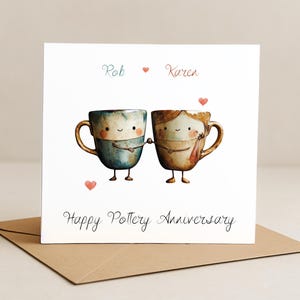 May include: A square greeting card featuring a watercolour illustration of two cartoon mugs holding hands. The mugs are blue and brown with smiling faces. The card reads "Rob Karen" and "Happy Pottery Anniversary."