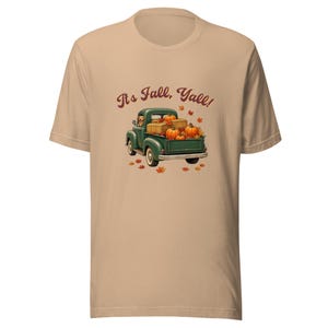 May include: A tan short-sleeve t-shirt featuring a graphic of a green vintage pickup truck carrying hay bales and pumpkins. A dog is visible in the driver's seat. The text "It's Fall, Yall!" is printed above the truck, surrounded by orange autumn leaves.