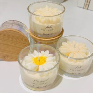 May include: Clear glass votive candles with daisy-shaped wax tops. One candle has a wooden lid. The candles have text that reads "Will you be my Bridesmaid?" and a name. The candles are on a white surface.