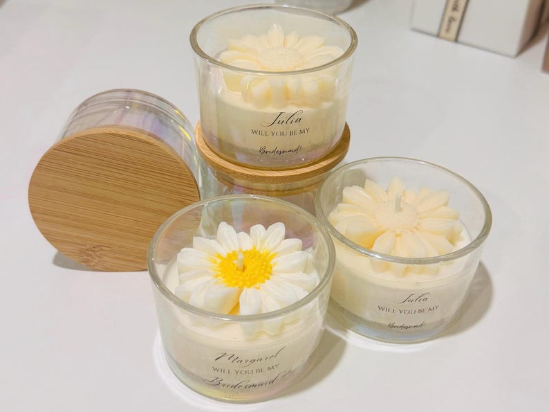 May include: Clear glass votive candles with daisy-shaped wax tops. One candle has a wooden lid. The candles have text that reads "Will you be my Bridesmaid?" and a name. The candles are on a white surface.
