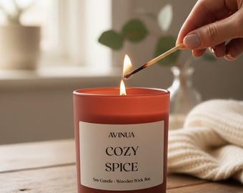 May include: A lit orange candle with the text "COZY SPICE" on a white label. A match is being used to light the candle. The candle is on a wooden table.