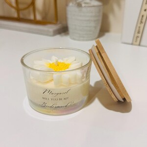 May include: A clear glass candle with a white daisy flower design on top and a wooden lid. The candle has text that reads "Margaret, Will You Be My Bridesmaid?" A decorative item for a wedding.