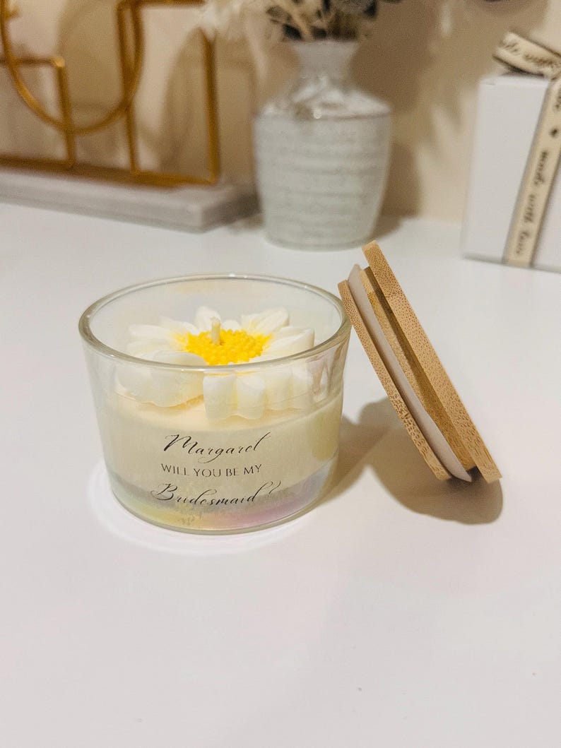 May include: A clear glass candle with a white daisy flower design on top and a wooden lid. The candle has text that reads "Margaret, Will You Be My Bridesmaid?" A decorative item for a wedding.