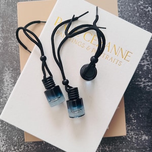 May include: Two small, clear glass bottles with a gradient blue tint, connected by black cords and beads. The bottles have black screw-on caps. The cords are attached to a black disc. The bottles are resting on a white book with gold lettering.