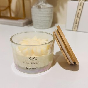 May include: A clear glass candle holder with a floral-shaped candle inside. The candle has a light yellow color. The candle holder has the text "Julia, Will You Be My Bridesmaid!" printed on it. A wooden lid is next to the candle holder.