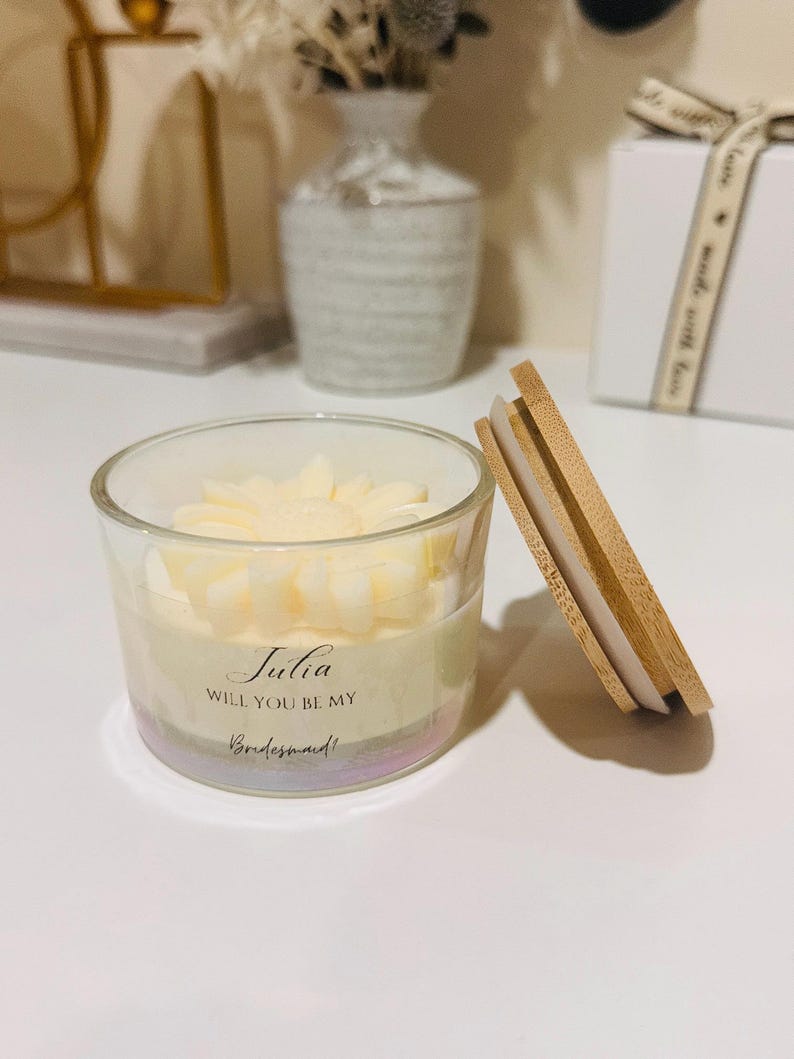 May include: A clear glass candle holder with a floral-shaped candle inside. The candle has a light yellow color. The candle holder has the text "Julia, Will You Be My Bridesmaid!" printed on it. A wooden lid is next to the candle holder.