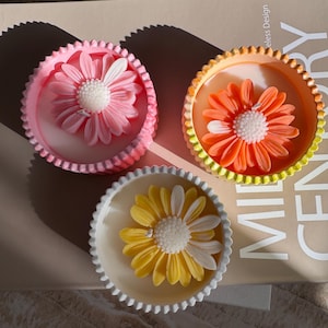 May include: Three decorative flower-shaped soaps in pink, orange, and yellow, each nestled in a scalloped dish. The soaps have white centers and are arranged on a neutral-toned surface, creating a vibrant and artistic display.