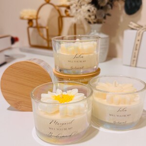 May include: Clear glass candles with floral designs and the text "Julia, Will You Be My Bridesmaid!" and "Margaret, Will You Be My Bridesmaid!" The candles have a white and yellow flower design on top. The candles sit on a wooden surface.