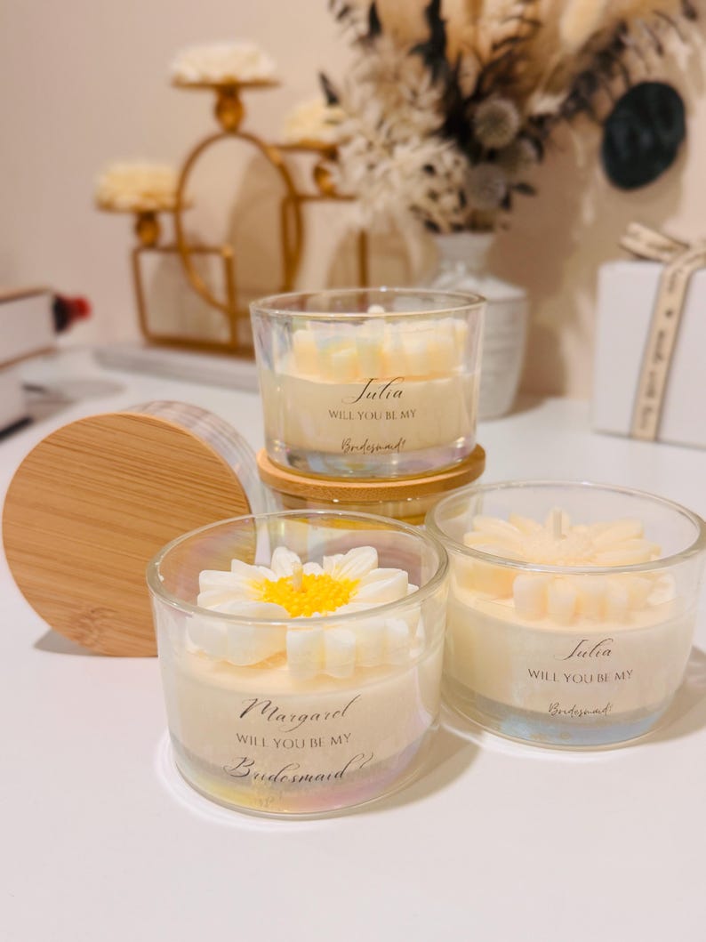 May include: Clear glass candles with floral designs and the text "Julia, Will You Be My Bridesmaid!" and "Margaret, Will You Be My Bridesmaid!" The candles have a white and yellow flower design on top. The candles sit on a wooden surface.