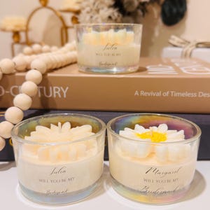 May include: Three clear glass candles with a floral design on top. The candles have a white wax base and are labeled with names and the text "Will you be my Bridesmaid?". The candles are on a table with decorative items.