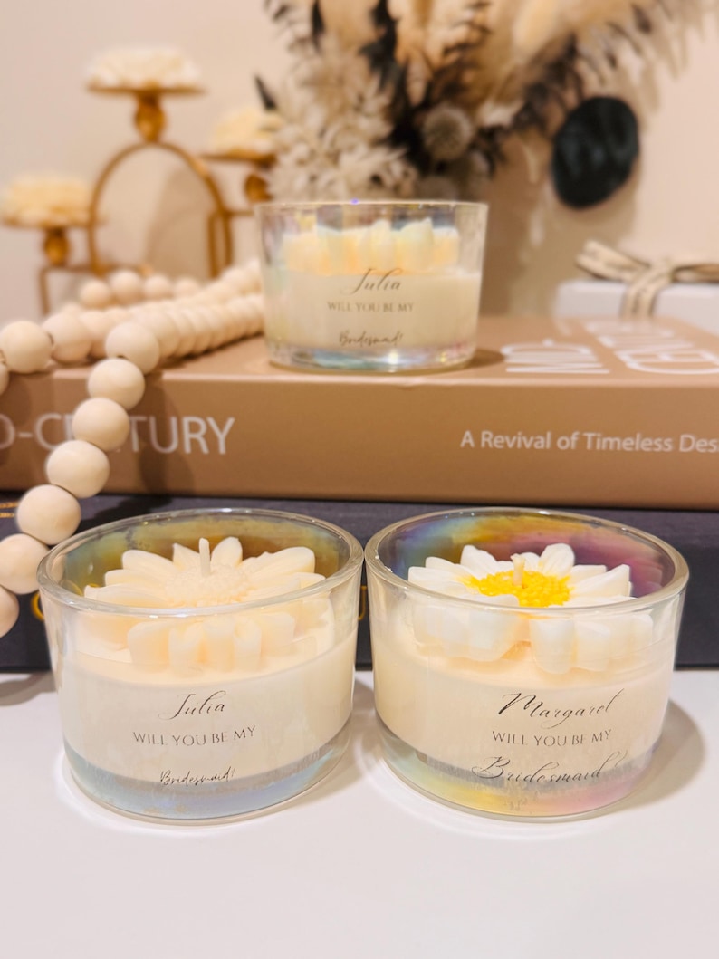 May include: Three clear glass candles with a floral design on top. The candles have a white wax base and are labeled with names and the text "Will you be my Bridesmaid?". The candles are on a table with decorative items.