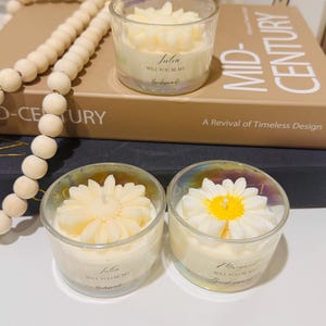 May include: Two glass votive candles with floral designs. One candle has a daisy design with a yellow center, the other has a flower design. The candles have text that reads "Julia, Will you be my Bridesmaid!" and "Margaret, Will you be my Bridesmaid!"