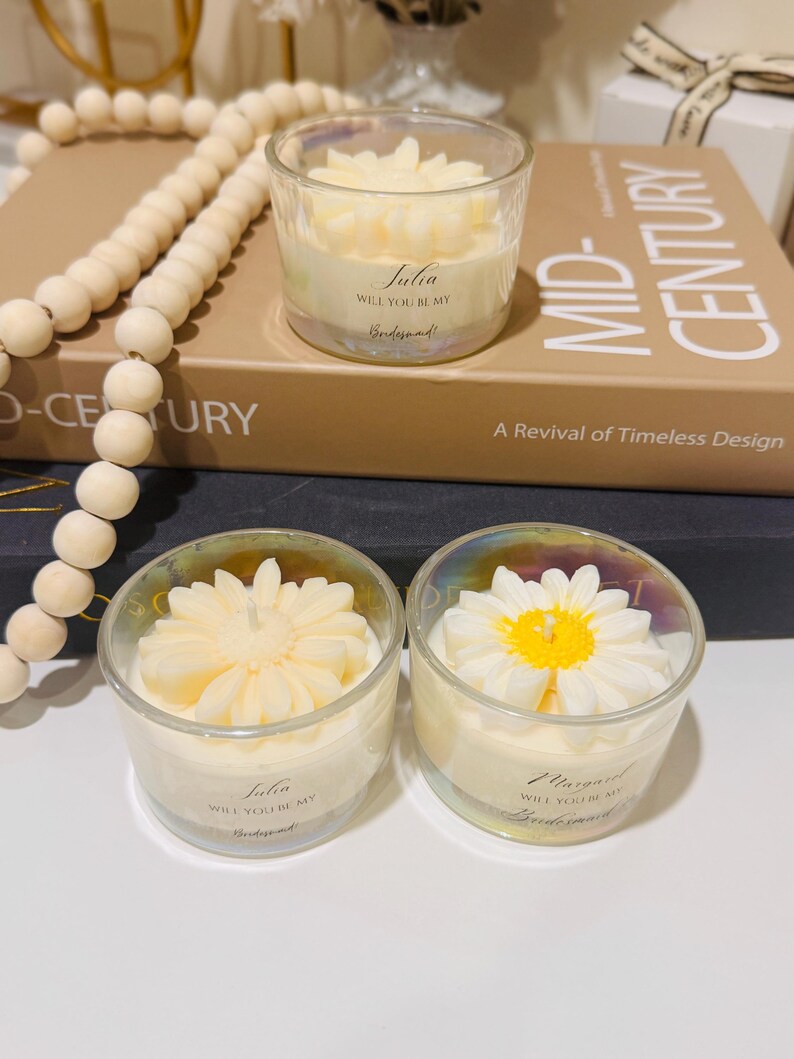 May include: Two glass votive candles with floral designs. One candle has a daisy design with a yellow center, the other has a flower design. The candles have text that reads "Julia, Will you be my Bridesmaid!" and "Margaret, Will you be my Bridesmaid!"
