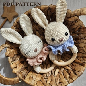 Può includere: Due giocattoli sonagli a forma di coniglio all'uncinetto con anelli di legno. Un coniglio indossa un colletto a balze rosa e l'altro un colletto a balze blu. I conigli sono in un cesto di vimini.