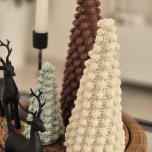 May include: Three crocheted Christmas trees in different sizes and colors. The largest tree is brown, the middle tree is green, and the smallest tree is white. The trees are sitting on a wooden tray.
