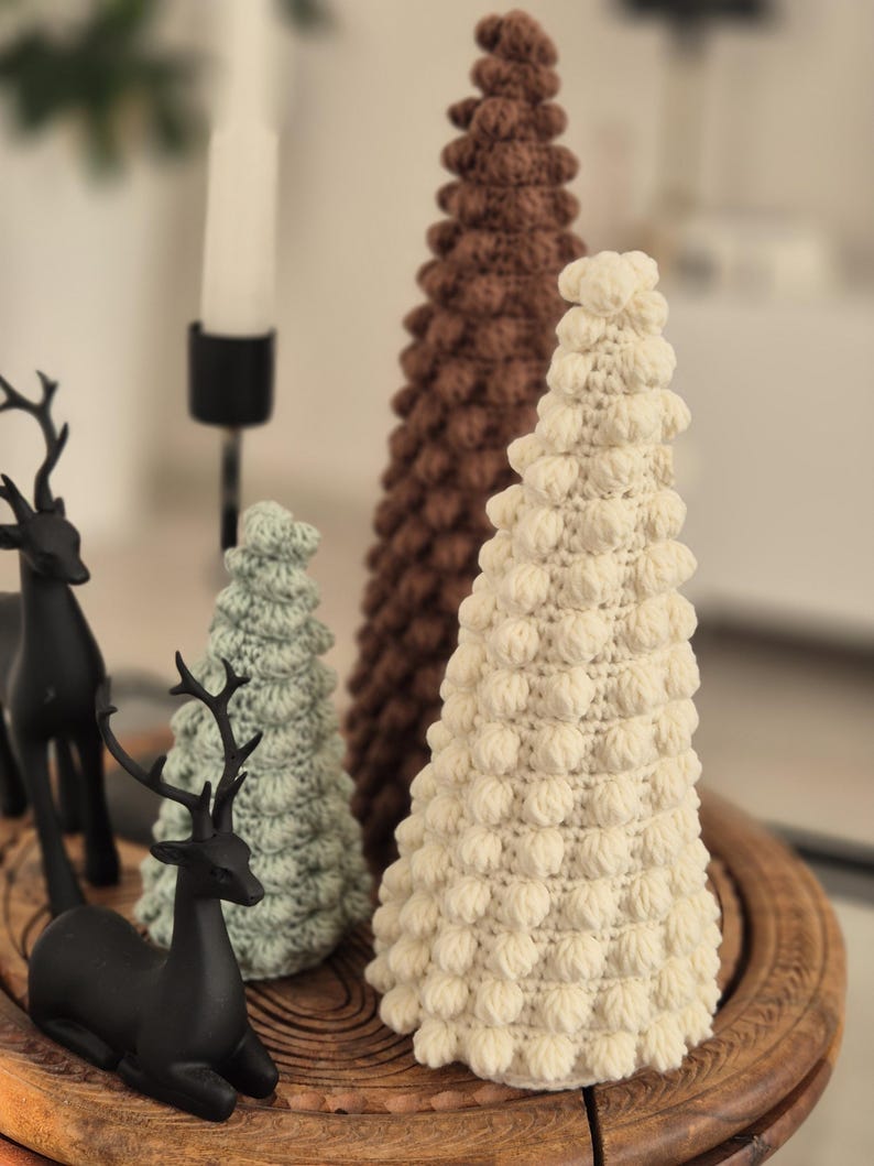 May include: Three crocheted Christmas trees in different sizes and colors. The largest tree is brown, the middle tree is green, and the smallest tree is white. The trees are sitting on a wooden tray.