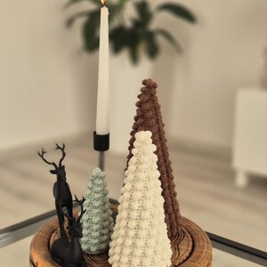 May include: Three crocheted Christmas trees in brown, white, and green on a wooden tray with two black reindeer figurines. A lit white candle is in the background.