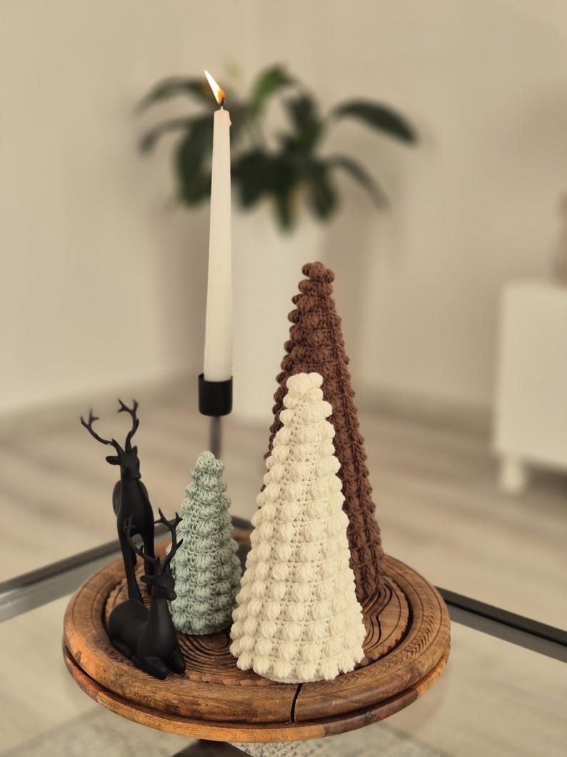 May include: Three crocheted Christmas trees in brown, white, and green on a wooden tray with two black reindeer figurines. A lit white candle is in the background.