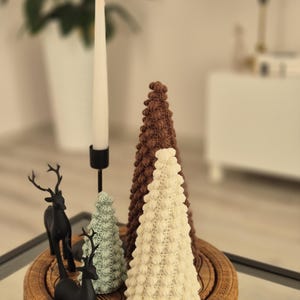 May include: Three crocheted Christmas trees in brown, white, and green on a wooden stand with two black reindeer figurines. A lit candle sits on a black candlestick.