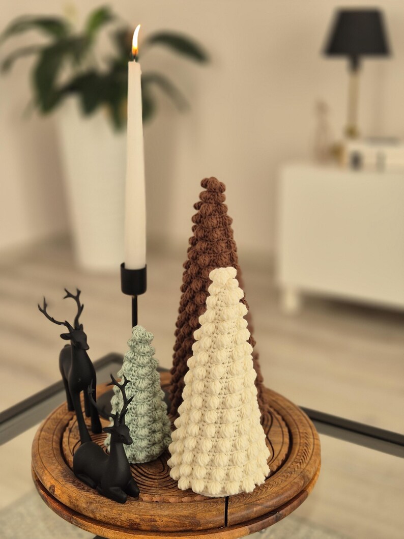 May include: Three crocheted Christmas trees in brown, white, and green on a wooden stand with two black reindeer figurines. A lit candle sits on a black candlestick.