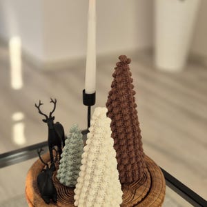 May include: A wooden tray with three crocheted Christmas trees and a black metal candle holder with a white candle. The trees are brown, white, and green.