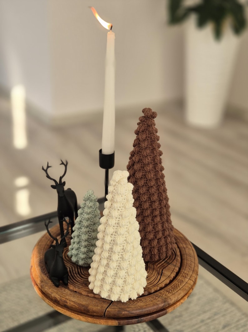 May include: A wooden tray with three crocheted Christmas trees and a black metal candle holder with a white candle. The trees are brown, white, and green.