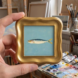 May include: A small square painting of a single sardine centered on a light blue background, encased in a thick, ornate, metallic gold frame with wavy edges. The fish is rendered in shades of silver, blue, and yellow with visible brushstrokes.