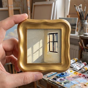 Puede incluir: Una pequeña pintura cuadrada en un marco dorado representa una ventana con luz solar. La obra de arte presenta una paleta de colores neutros con juegos de luces y sombras, creando una estética minimalista. La pintura se sostiene en una mano.