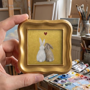 May include: A small square painting in a decorative gold frame featuring a white rabbit and a brown rabbit sitting side by side against a textured gold background. A small red heart is painted above the rabbits, centered in the upper portion.