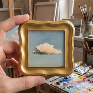 May include: A small square oil painting featuring a single soft pink cloud centred against a pale blue sky, encased in a decorative gold-toned frame with a wavy, scalloped edge. The artwork depicts a serene atmospheric scene with visible brushstrokes.