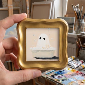 May include: A small square painting in a gold ornate frame featuring a white ghost with black eyes sitting in a white bathtub filled with bubbles. The background is a soft beige color with several floating bubbles surrounding the ghost.