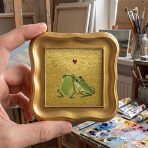 May include: A small square painting in a gold ornate frame featuring two green frogs sitting side by side on a textured gold background with a single small red heart painted above them. The artwork is held up to display the detailed brushwork.