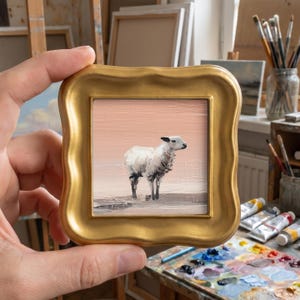 May include: A small square oil painting of a white sheep standing in a field, rendered with visible brushstrokes against a soft peach-coloured sky. The artwork is encased in a decorative, ornate gold-toned frame with a wavy, scalloped edge.