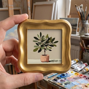 May include: A small square painting of a potted olive tree with green leaves and dark fruit, set in a decorative gold frame with wavy edges. The plant sits in a light pink pot against a neutral beige background with a soft shadow cast below.