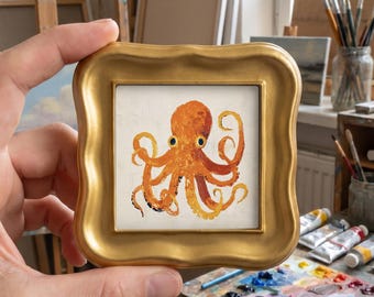 May include: A small square painting of an orange octopus with dark eyes, centered on a light background. The artwork is encased in a thick, wavy-edged gold frame with a smooth, metallic finish, highlighting the detailed brushstrokes of the creature.