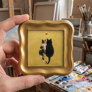 May include: A small square painting in a gold ornate frame featuring a calico cat and a black cat sitting side by side against a textured gold background. A tiny red heart is painted above the two cats, who are depicted in a thick impasto style.