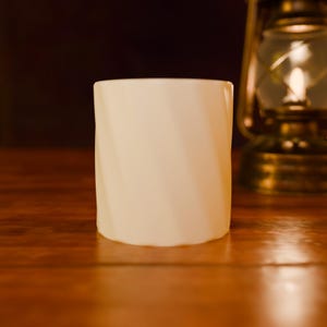 Könnte beinhalten: Ein zylindrischer weißer Becher mit einer subtilen, diagonal verlaufenden Spiral- oder Drehstruktur auf der Außenfläche. Der Becher hat eine glatte, matte Oberfläche und ein schlichtes Design, steht aufrecht mit einem flachen Boden und einem breiten Rand.