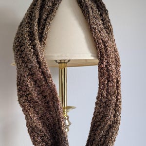May include: A brown and gold infinity scarf made of a soft, textured yarn. The scarf is draped over a lamp.