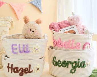 May include: Four off-white rope baskets with embroidered names and floral accents. The baskets contain a stuffed bear, a pink unicorn, a pink blanket, and a book. The names are "Ell", "Hazel", "Isabelle", and "Georgia".