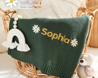 May include: A dark green knitted baby blanket with the name "Sophia" embroidered in gold, adorned with daisy accents. A white rainbow-shaped toy with tassels and a teddy bear are next to the blanket, which is displayed in a wicker basket.