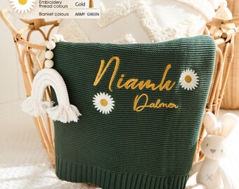 May include: Army green knitted baby blanket with gold embroidered name and daisy accents. A white crochet rainbow and a stuffed bunny are attached to a wicker basket. The blanket is personalized with the name "Niamh Dalmer".