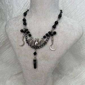 May include: A gothic necklace with a bat pendant, crescent moon charms, and a black crystal. The necklace is made with black beads and silver-toned metal accents. The design is displayed on a white mannequin bust.