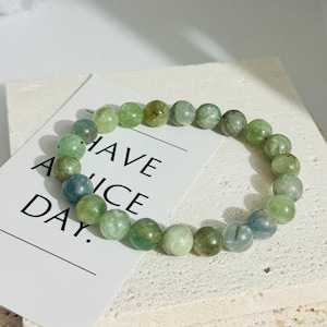 Peut inclure: Un bracelet en pierres précieuses vertes avec des perles rondes. Le bracelet est sur une surface blanche avec une carte qui dit "Have a nice day."
