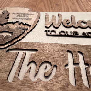 May include: A wooden sign with the text "Welcome to our adventure The" and a National Park Service logo featuring a tree and a bison. The sign is made from two different types of wood, one light and one dark.