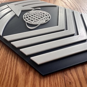 May include: A white 3D printed emblem with a globe and arrow design on a dark blue background. The emblem is shaped like a hexagon.