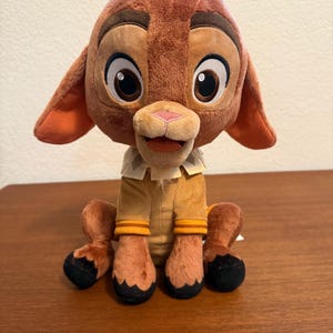 May include: A plush toy of a brown fawn with large dark eyes, a pink nose, and a small tuft of hair on its head. The fawn wears a tan shirt with orange striped cuffs and a white collar with a geometric pattern. It has black hooves and sits upright.