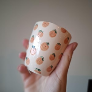 Könnte beinhalten: Eine weiße Keramiktasse mit einem handgemalten Orangemuster. Die Orangen sind in Pfirsich- und Orangetönen mit grünen Blättern gehalten. Die Tasse wird in einer Hand gehalten, um das Design und die Form zu zeigen.