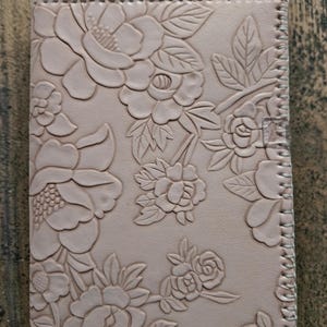 May include: A rectangular light pink leather notebook cover featuring an embossed floral pattern of various flowers and leaves. The edges are finished with a visible whipstitch detail using light-colored thread, creating a textured border.